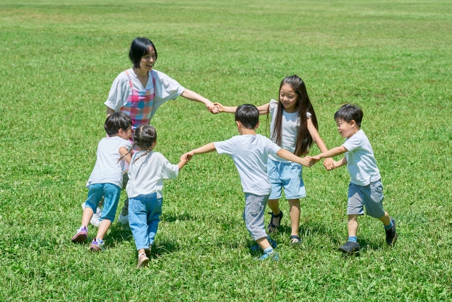 芝生の上で手を繋いで輪をつくり遊ぶ子ども達と女性